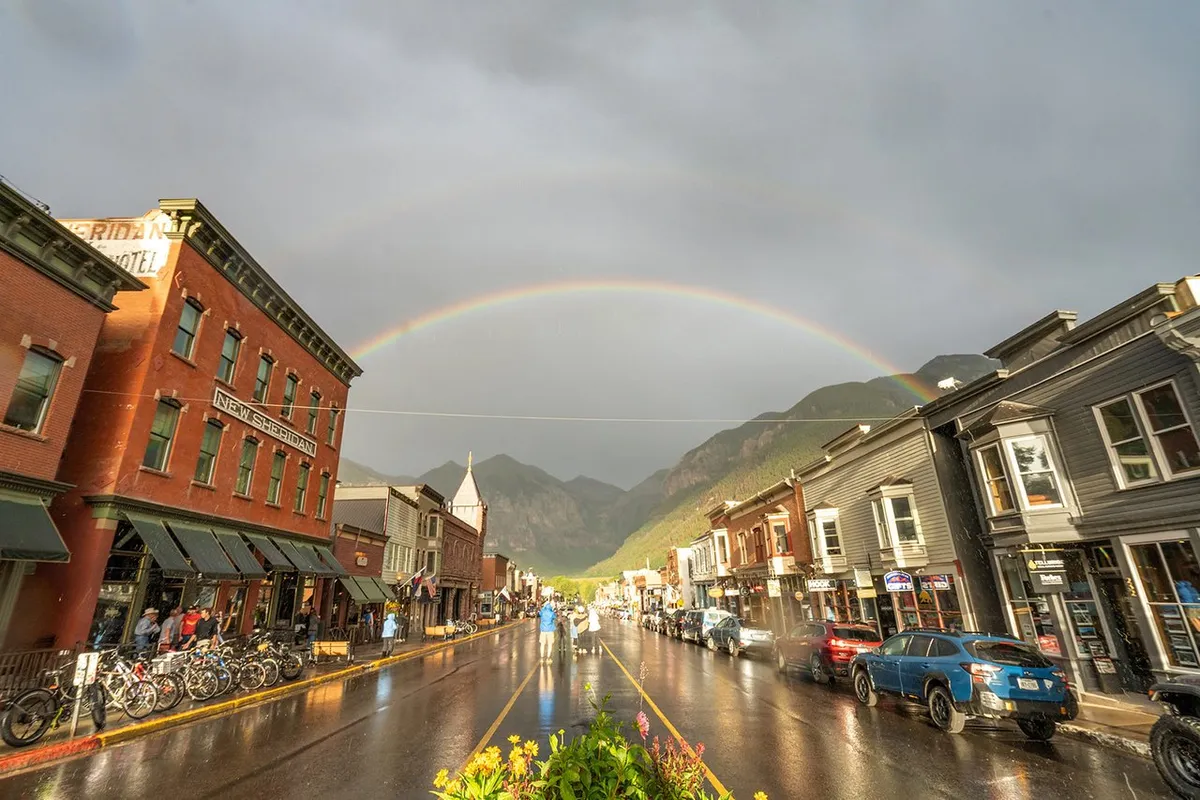 This image captures Telluride’s Main Street bustling with activity. People are seen walking and biking along a street lined with colorful historic buildings. Bicycles rest near the sidewalk, and a rainbow arches gently over the town, evoking a lively yet charming small-town vibe. Outdoor cafe tables hint at local dining, and the scene feels fresh, light, and welcoming.