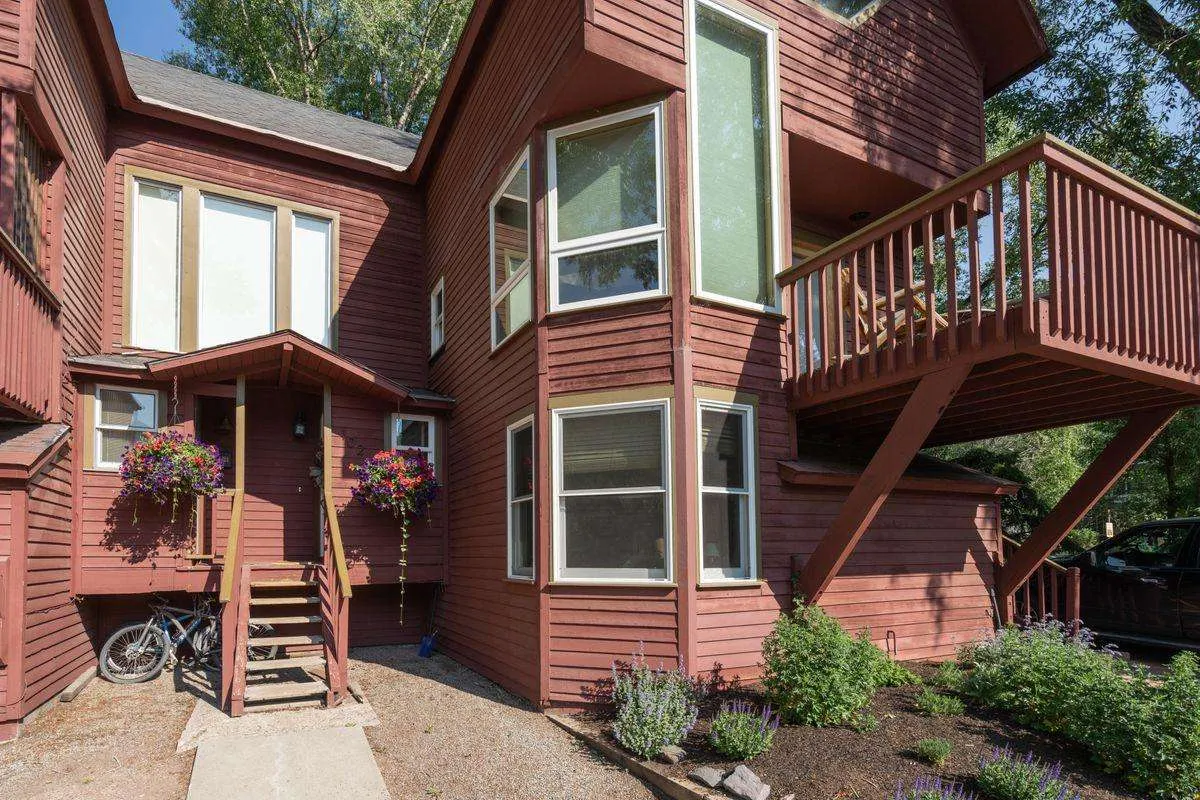 A welcoming exterior shot of the building's entrance, featuring a staircase, balcony with people gathering, flower pots, and large windows. The scene conveys a sunny, safe, and inviting neighborhood setting.