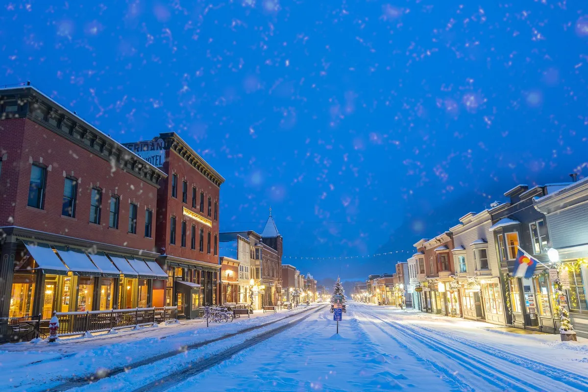 A snow-dusted historic street in Telluride lined with festive lights, a decorated Christmas tree, and warmly lit shops. Streetlights and softly falling snow create a cozy, whimsical winter village atmosphere.