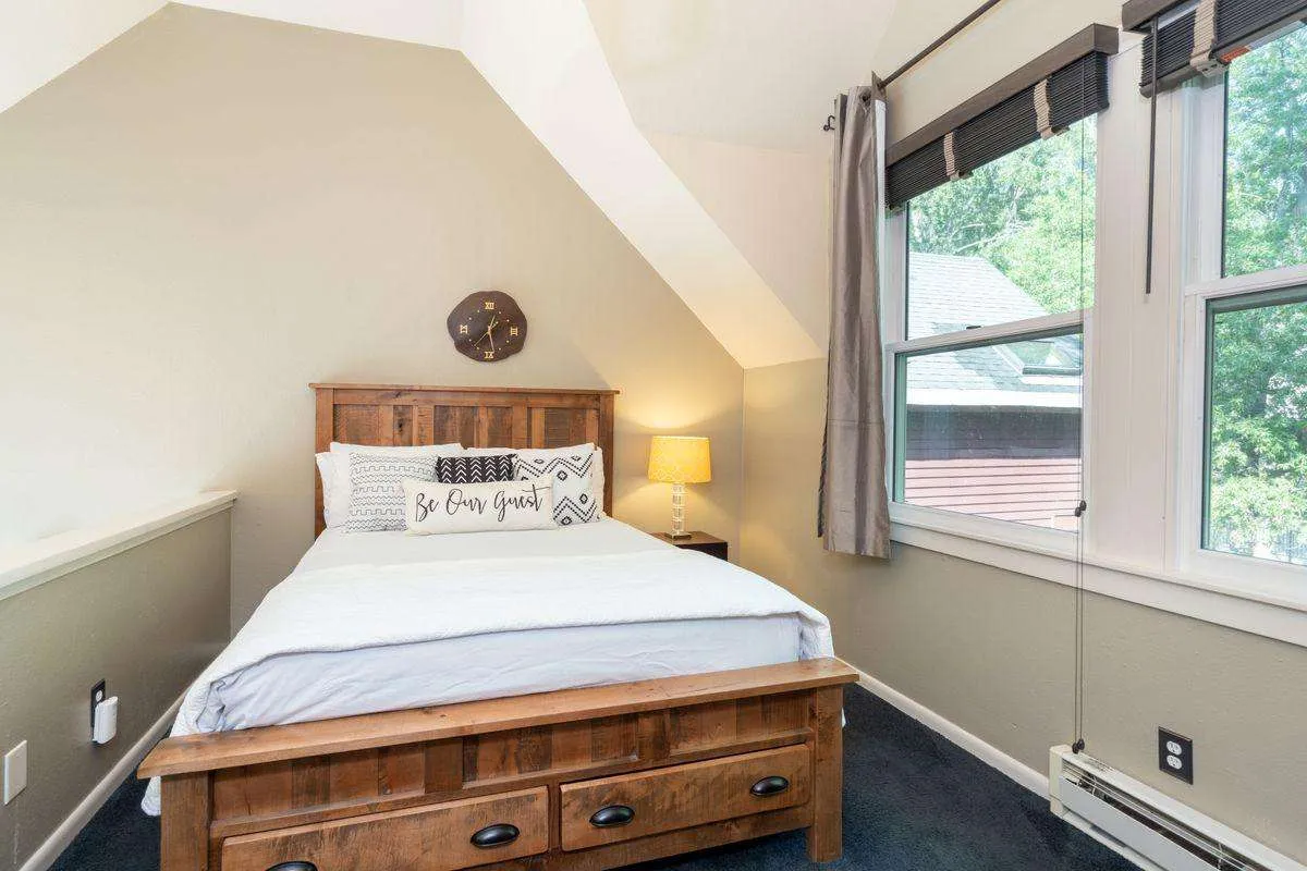 A cozy, bright lofted bedroom space with a wooden queen bed, plush decorative pillows, a bedside lamp for nighttime reading, and large windows that let in natural light and offer views of the surrounding landscape. The ambiance is intimate and inviting, perfect for relaxing or enjoying quiet mornings.