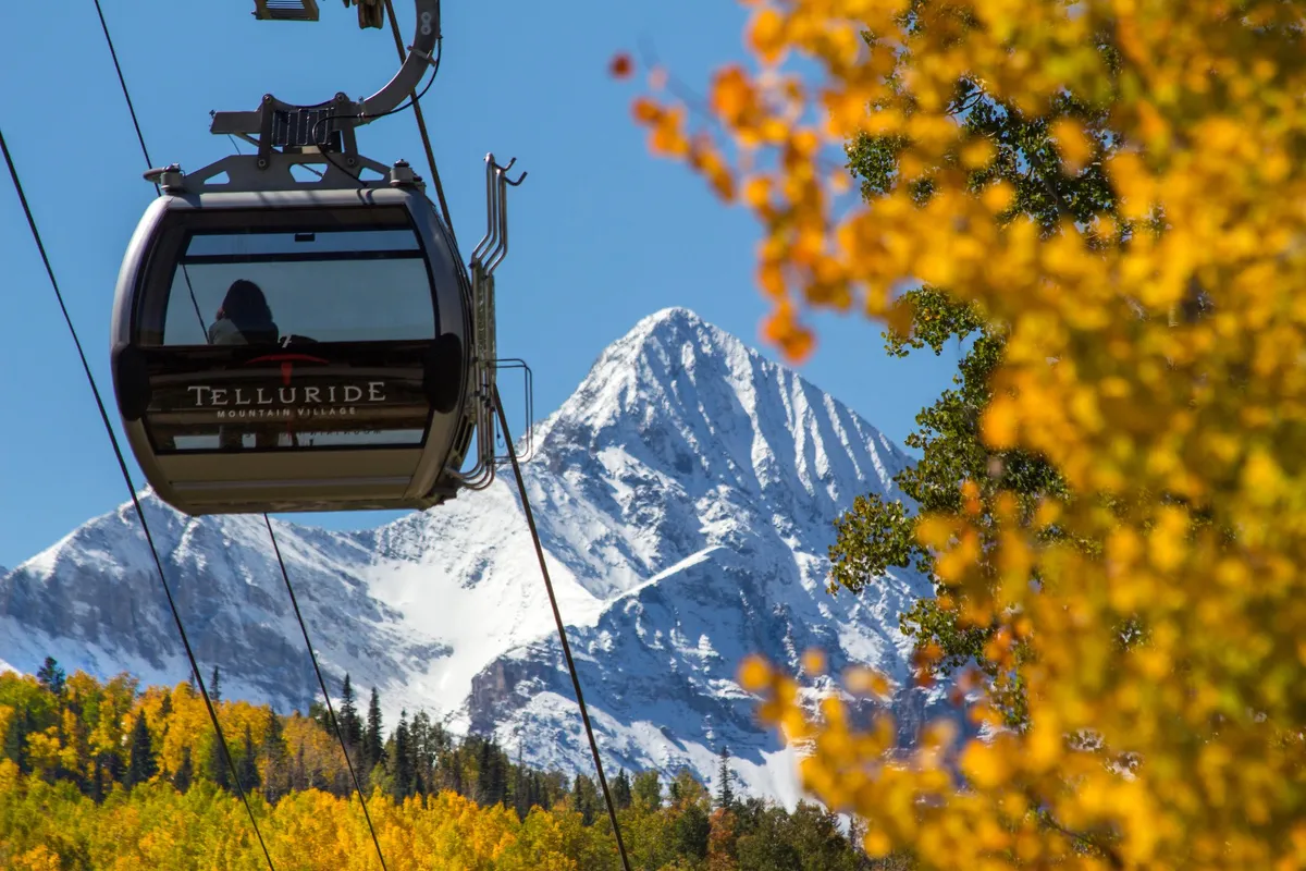 A gondola car glides above a snow-capped mountain, framed by vibrant fall foliage in the foreground. The scene captures both the thrill of scenic rides and the proximity to mountain adventures, with the iconic Telluride landscape visible in the background.