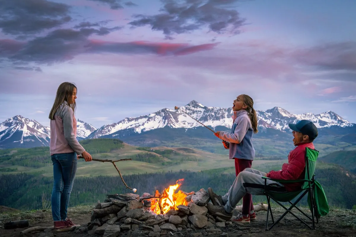 A picturesque outdoor scene featuring a campfire surrounded by children roasting marshmallows, an adult sitting calmly nearby, and a stunning mountain landscape in the background. The atmosphere is warm and joyful, emphasizing the connection between families, children, and nature.