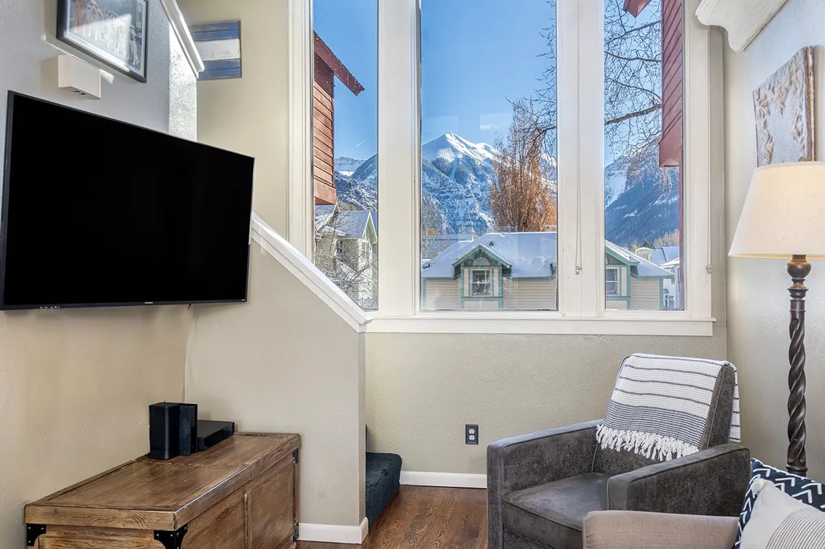 A spacious, light-filled living room featuring large windows with mountain views, a comfortable armchair, a flat-screen TV, and a wooden coffee table. The ambiance is both warm and inviting, with plenty of natural light and cozy furnishings that invite relaxation and conversation.
