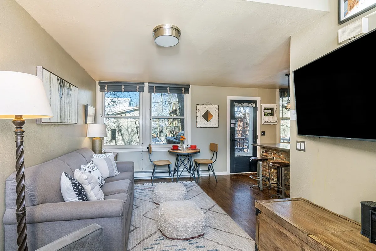 An open-plan living and dining area featuring a comfortable sofa, dining table, television, and stylish lighting. Expansive windows provide plenty of daylight, enhancing the cozy and welcoming mood.